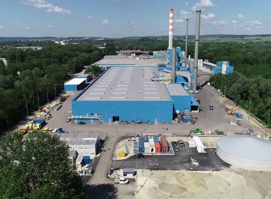 Luftaufnahme eines großen Industriekomplexes mit blauen Produktionshallen, hohen Schornsteinen und angrenzenden Betriebsflächen; das Werk liegt inmitten von Grünflächen und Wald unter klarem Himmel.